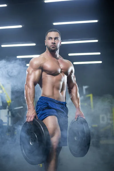 Handsome muscular athlete man is standing in a gym against the background of smoke holding barbell discs in both hands — Stock Photo, Image