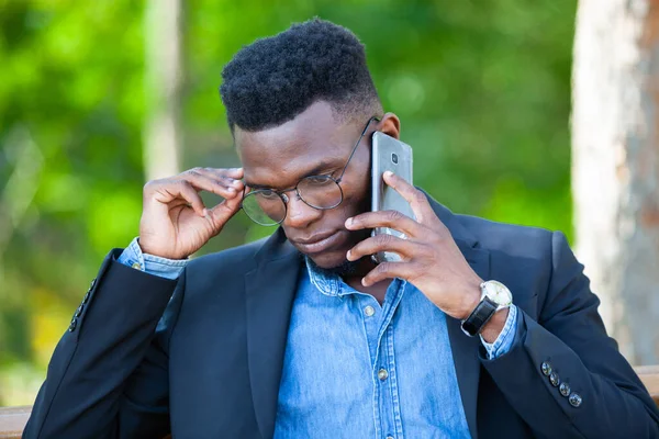 Portré egy magabiztos afrikai üzletemberről, aki beállítja a szemüvegét és telefonon beszél egy nyári parkban. — Stock Fotó