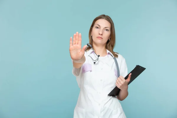 La hermosa doctora con un estetoscopio y una tableta negra, muestra una señal de stop con la mano, sobre un fondo azul. Copiar pasta. concepto de salud. Vamos a detener la enfermedad. —  Fotos de Stock