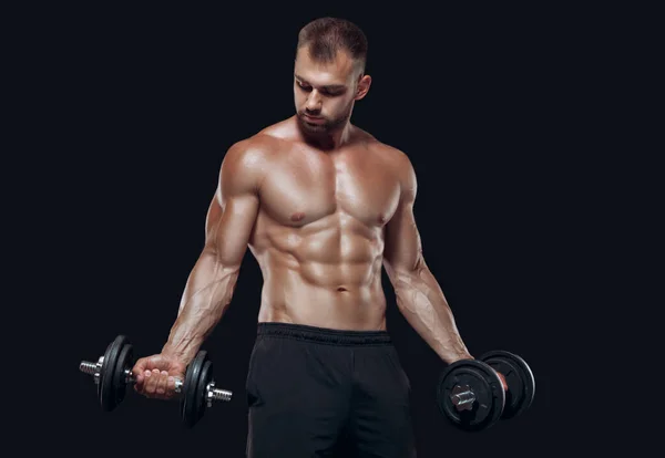 性感的运动员表现出肌肉发达的身体，低着头站着哑铃，与黑色背景隔离 — 图库照片