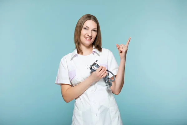 Seorang dokter wanita cantik dengan stetoskop menunjuk ke tempat di mana akan ada informasi penting, di latar belakang biru. Salin pasta. konsep layanan kesehatan. — Stok Foto