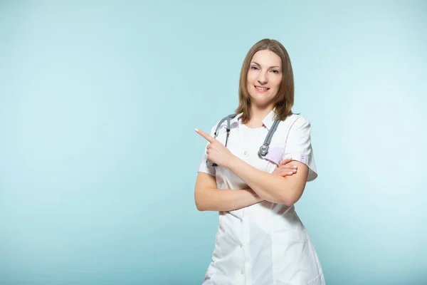 A beautiful female doctor with a stethoscope points to the place where there will be important information, on a blue background. Copy paste. healthcare concept. — Stock Photo, Image