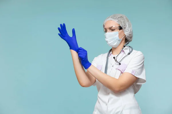 Se pone guantes azules. Médico o enfermera con traje de protección personal con estetoscopio, sobre fondo azul. En una máscara, guantes, gafas y un sombrero. Copiar pasta. concepto de salud. —  Fotos de Stock