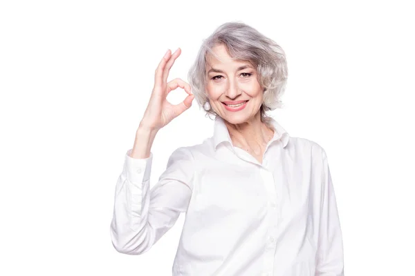 一个成熟迷人的女人穿着雅致的衬衫站在孤零零的白色背景上，笑容可亲地做着一个不错的手势的画像 图库图片