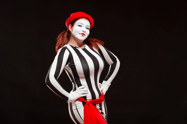 Portrait of female mime artist performing, isolated on black background. Woman stands with her hands on her sides, posing in striped dress smiling Stock Photo