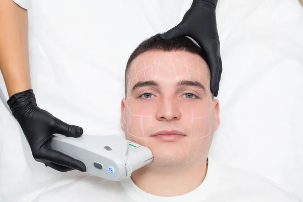 Portrait d'un jeune homme couché sur le canapé d'un bureau de cosmétologie avec des marques sur son visage appliquées avec un crayon cosmétique blanc et la main d'un cosmétologue dans un gant médical effectuant une procédure Images De Stock Libres De Droits