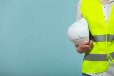Yakın plan. Profesyonel mühendis. Parlak yelek giymiş bir kadın mavi arka planda elinde koruyucu bir miğfer tutuyor. Yapıştırıcı kopyala.