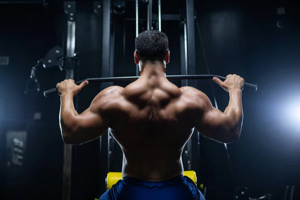 Handsome fitness man is performing back workouts using thrust of the upper block machine in a gym, back view — Stock Photo, Image