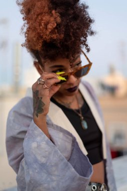 İspanya 'nın Cadiz kentindeki bulutsuz mavi gökyüzüne karşı terasta dinlenirken leylak bornoz takan kıvırcık saçlı ve güneş gözlüklü şık siyah kadın..