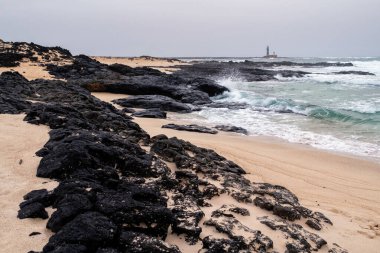 Soğuk bir günde, kumsaldaki deniz fenerinin gri gökyüzüne bakan uzak görüntüsü.