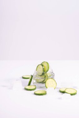 Beyaz zemin üzerinde buz küpleri olan cam kabın içinde ferahlatıcı kozmetik salatalıklar.