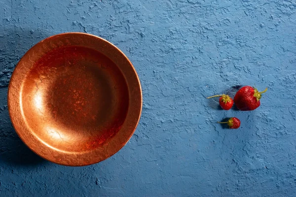 minimalistic still life with a copper bowl and three strawberries on a blue textured background. creative concept.