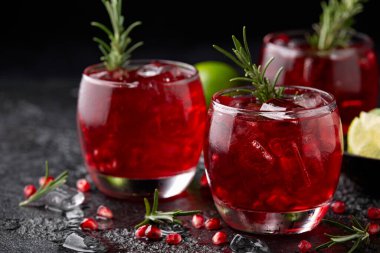 Pomegranate gin cocktail with lime, rosemary and ice on rustic black table.