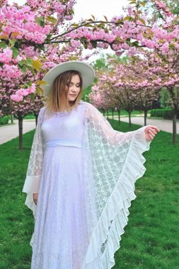 A pregnant woman in a white dress and hat surrounded by sakura.