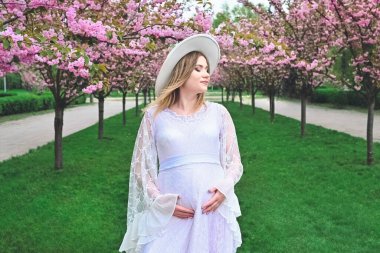 A pregnant woman in a white dress and hat surrounded by sakura.