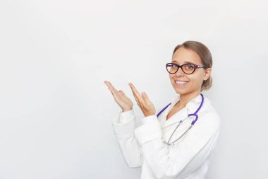 Beyaz önlüklü, gözlüklü, boynunda steteskop olan genç sarışın bir kadın doktor, beyaz arka planda elleri izole bir şekilde metin veya tasarım için boş bir kopya alanı gösteriyor.