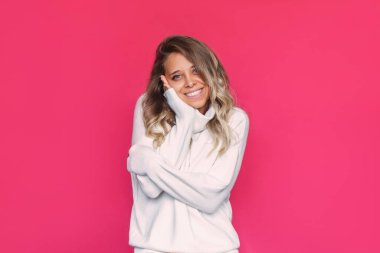 Beyaz yünlü, sıcak kaşmir kazaklı, dalgalı saçları olan beyaz, çekici bir sarışın kadın, parlak pembe bir arka planda kendini kucaklayan kumaşın yumuşaklığından zevk alıyor.