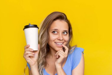 Genç, düşünceli, sarışın bir kadının elinde beyaz bir fincan kahve ya da çay tutan ve sarı arka planda izole edilmiş bir şekilde bakışları. Metin veya tasarım için kopyalama alanı ile modelleme