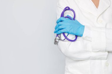 Beyaz önlüklü genç bir doktorun elinde steteskop ve gri arka planda izole edilmiş mavi bir tıbbi eldiven tutarken çekilmiş görüntüsü.