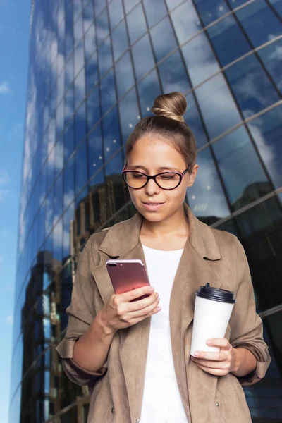 Young successful blonde business woman in office-style clothing and glasses holds a white paper cup of coffee or tea looking at a mobile phone screen on the background of a business center.