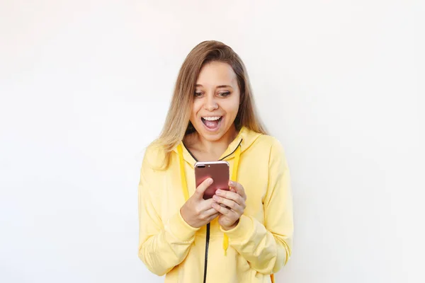 黄色のスポーツジャケットを着た若い白人の幸せなブロンドの女性が笑顔で 白い黄色の背景に隔離された画面を見て携帯電話を保持しています 驚いた女の子は彼女のスマートフォンを使用します — ストック写真