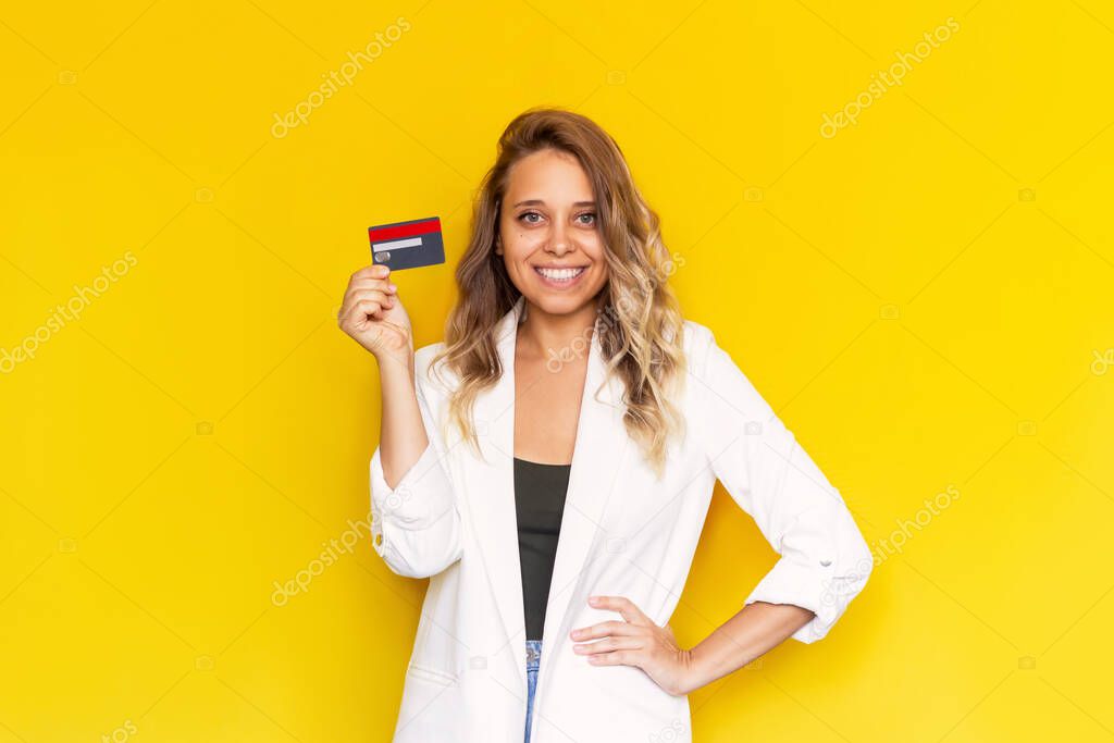 Smiling blonde woman in a white jacket holding a plastic credit card in her hand isolated on yellow background. Shopping, payment for purchases, banking operations