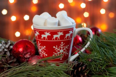 Geyik desenli kırmızı kupa, meşrubat desenli kar taneleri ve Noel ağacının dalında marşmelov, oyuncak toplar ve bulanık çelenk ışıklarının arka planında koni. Yeni yıl süslemeleri