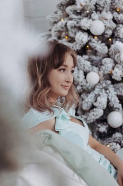 Beyaz bir Noel ağacının arka planında oturan neşeli elbiseli, beyaz, beyaz, çekici, gülümseyen bir kadın. Yeni Yıl İçi