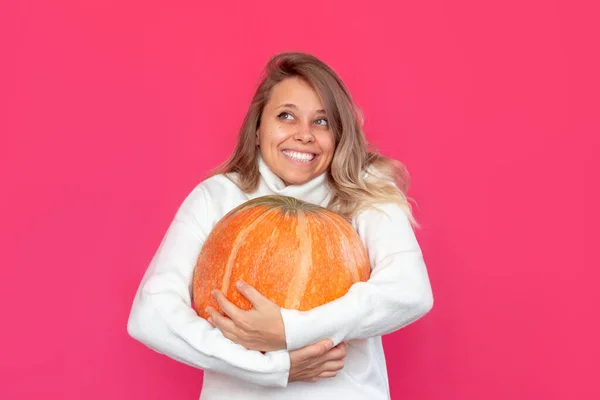 A young caucasian pretty smiling blonde woman in a white wool warm sweater holds a large pumpkin in her hands isolated on a bright color pink background. Halloween concept