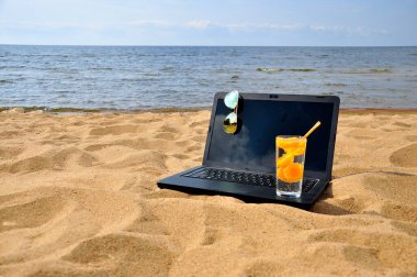 Kumsalda uzaktan kumanda, siyah bir laptop ve güneş gözlüğü..