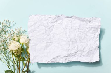 Buruşuk bir kağıt parçası ve bir buket beyaz gül. Üzerinde gölge ve açık mavi bir zemin var. Uzayı kopyala Düz yatış, üst görünüm.