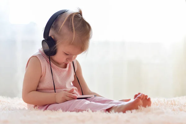 Una niña de dos años de edad está sentada en auriculares negros y viendo dibujos animados en su teléfono. —  Fotos de Stock