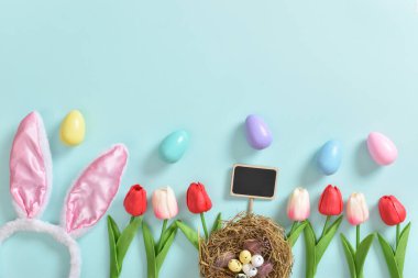 Üzerinde kartpostal ve hediye olan bir buket güzel lale ve açık mavi arka planda yumurtalar ve tüylerle dolu küçük bir sepet. Paskalya bayrağı. Kopyalamak için yer var. Düz konum, üst görünüm.