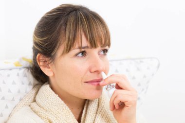 A young woman in bed suffering from a cold, with a nasal spray.