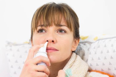 A young woman in bed suffering from a cold, with a nasal spray.