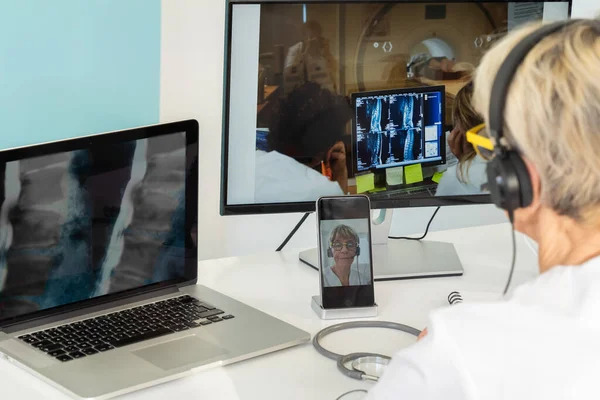 Teleconsultation between two doctors with medical images of the spine on one of the screens and team around a scanner on the other.