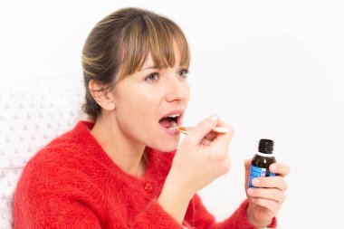 A young woman in bed suffering from a cold with cough syrup.
