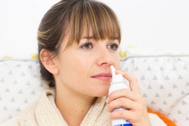 A young woman in bed suffering from a cold, with a nasal spray.
