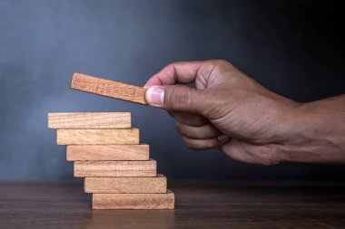 Yakın plan el, çöküşü veya finansal risk yönetimi ve stratejik planlama kavramlarını önlemek için piramit şeklinde dizilmiş ahşap blok kulesini ihtiyatlı bir şekilde yerleştiriyor.