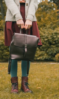 Örgü ceketli, gri taytlı ve etekli genç bir kadın. Şık bordo ayakkabılar. Kollarda deri çanta. Yeşil çimenler. Sonbahar modası.