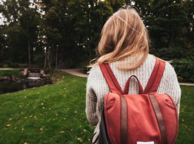 Örgü ceketli genç bir kızın arka görüntüsü. Havalı bir sırt çantası. Sonbahar modası. Şehir parkında yürürken.