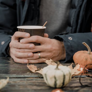 Kartondan bir fincan kahveyi adamın eline yakın çekim. Sonbahar sıcak içkisi. Balkabakları, akçaağaç yaprakları ve çelenkler eski ahşap masada.