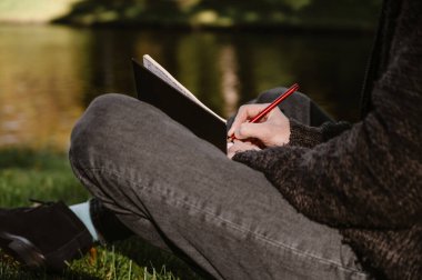 Öğrenci parkta çimlerin üzerinde oturuyor ve notlar alıyor. Arkaplanda gölet.