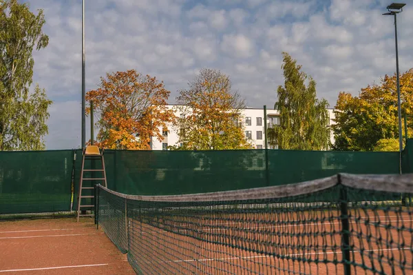 Empty outdoor clay tennis court at autumn cloudy day. Net and seat for referee. Green and yellow trees.