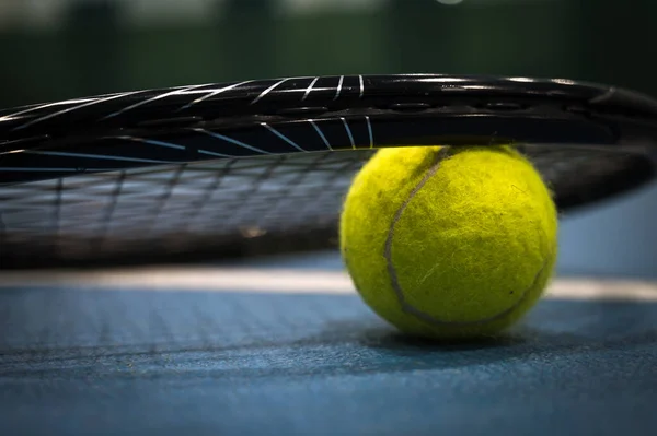 Close-up of yellow tenis ball under racket on blue hard tenis court.
