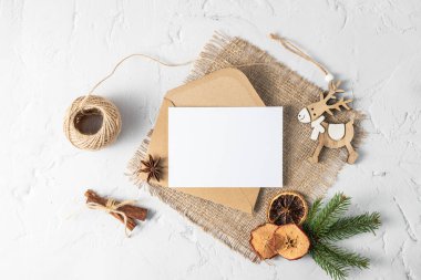 Köknar dalı, boş kağıt, zarf, ahşap geyik ve beyaz taş arka planda kış dekoru olan mutlu noel kutlaması kartları. Tarçın çubukları, anason yıldızı ve kurumuş meyveli Noel mektubu.