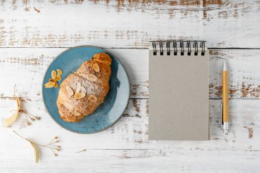Klasik mavi tabakta taze badem fıstıklı kahvaltı kruvasanı ve kırsal ahşap arka planda bambu kalemli boş defter defteri. Fotokopi alanı olan üst görünüm biçimlendirme pasta kartı