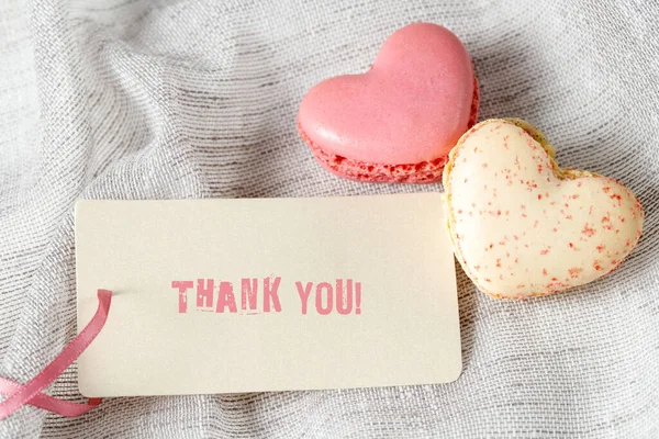 Thank you gift card with text and two macaroons in heart shape on light grey textile background. French cookies with raspberry and strawberry