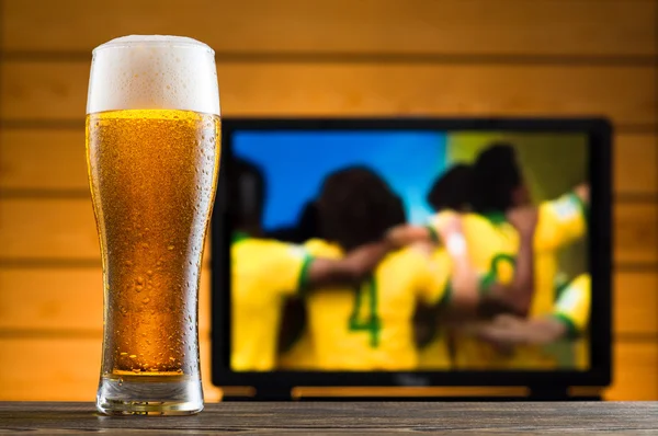 A glass of cold beer on the table, football match in background