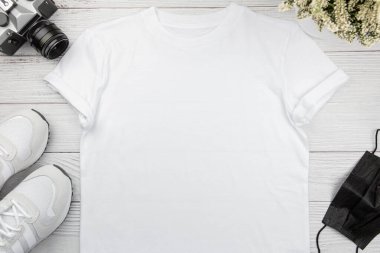 Beyaz kadın pamuk tişörtü, spor ayakkabısı, retro kamera ve beyaz tahta arka planda yüz maskesi. Tişört şablonu tasarla, baskı sunumunu düzenle. Üst görünüm düzlüğü.